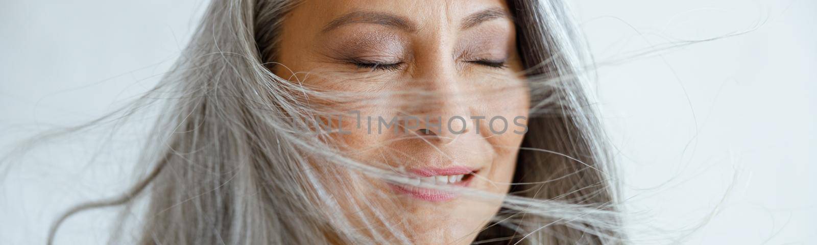 Happy Asian female model with flying hoary hair stands on light grey background in studio closeup. Mature beauty lifestyle