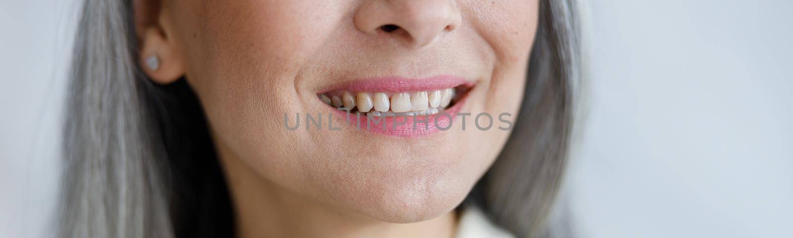 Positive mature lady in beige shirt smiles to camera showing healthy teath on light background in studio closeup