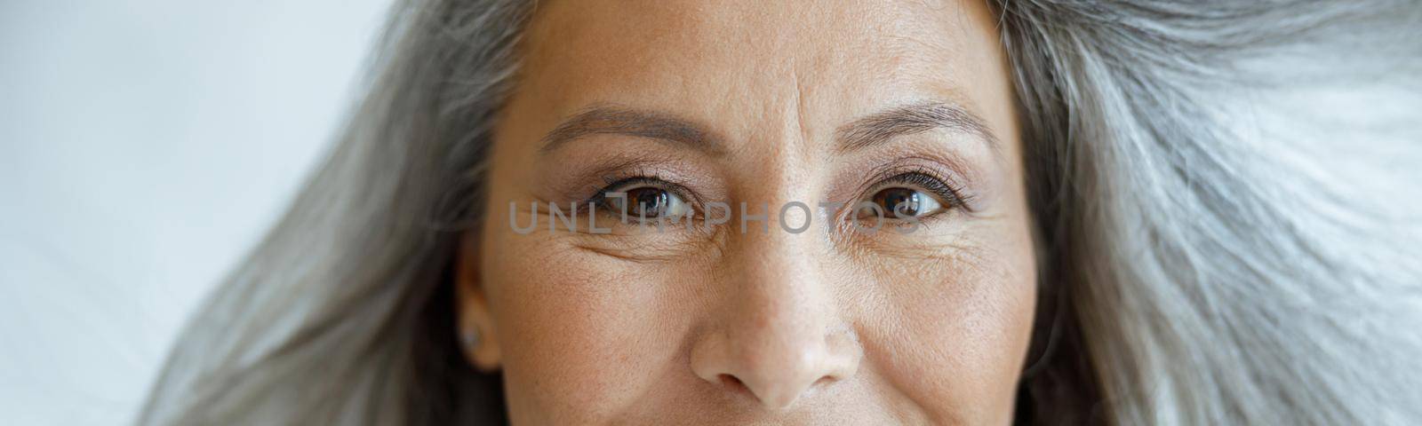 Positive middle aged Asian woman with flying hoary hair looks into camera on light grey background in studio closeup. Mature beauty lifestyle