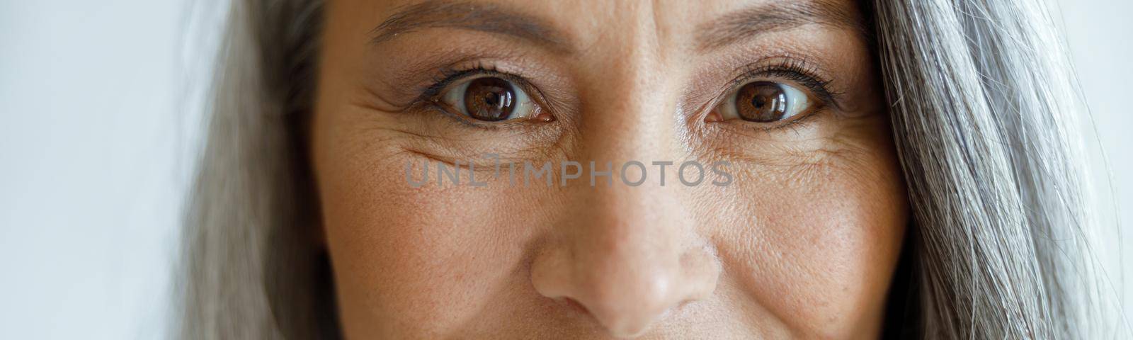 Cheerful silver haired lady with light makeup looks into camera on light grey background in studio closeup. Mature beauty lifestyle