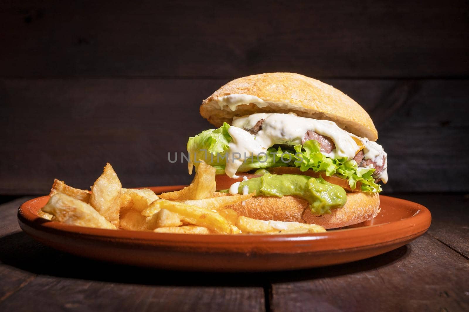 Delicious gourmet beef burger with creamy sauce, lettuce, tomato and guacamole, served with french fries on a rustic plate on a wooden table. by HERRAEZ