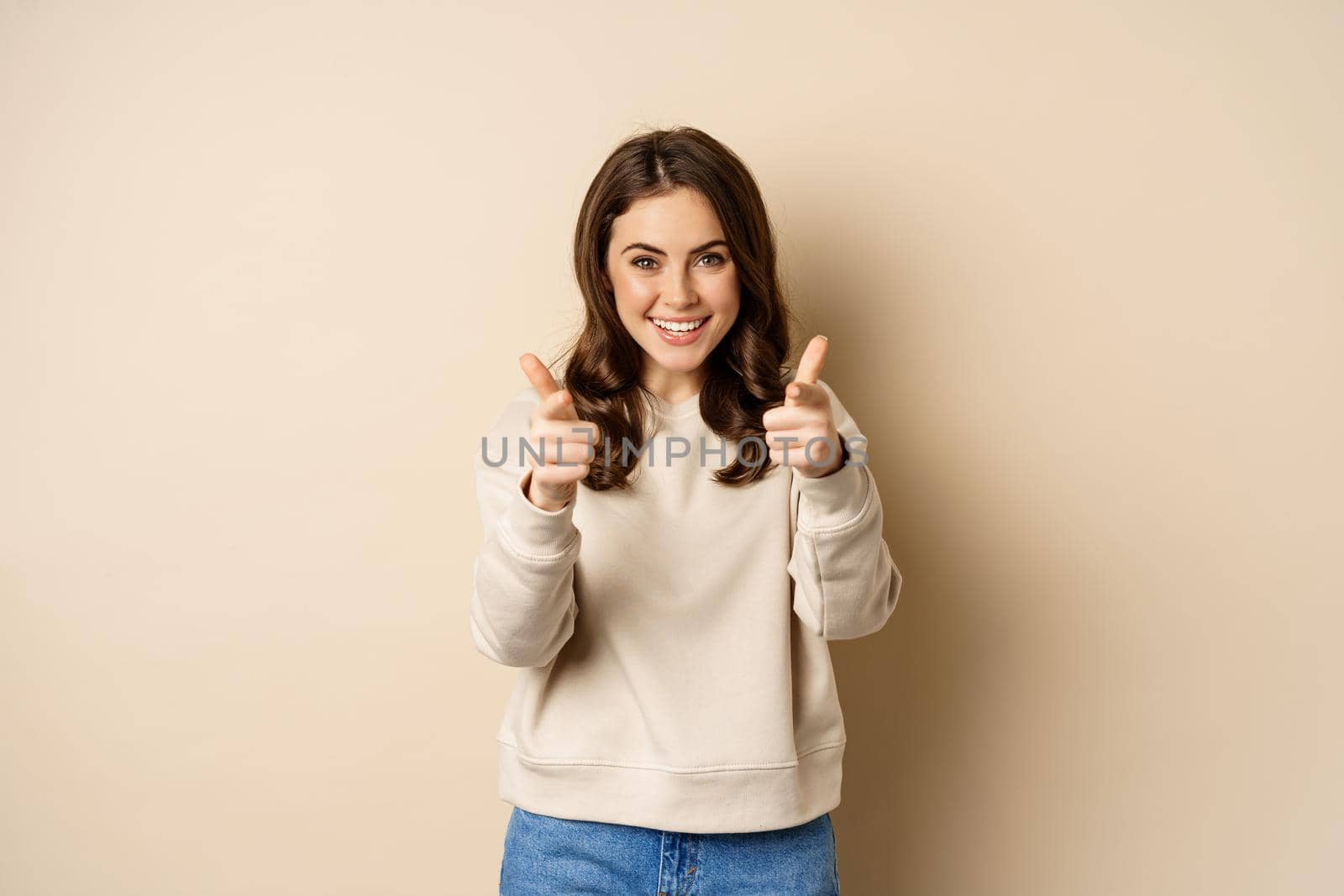 Smiling happy woman choosing you, pointing fingers at camera and laughing, inviting people, standing over beige background by Benzoix