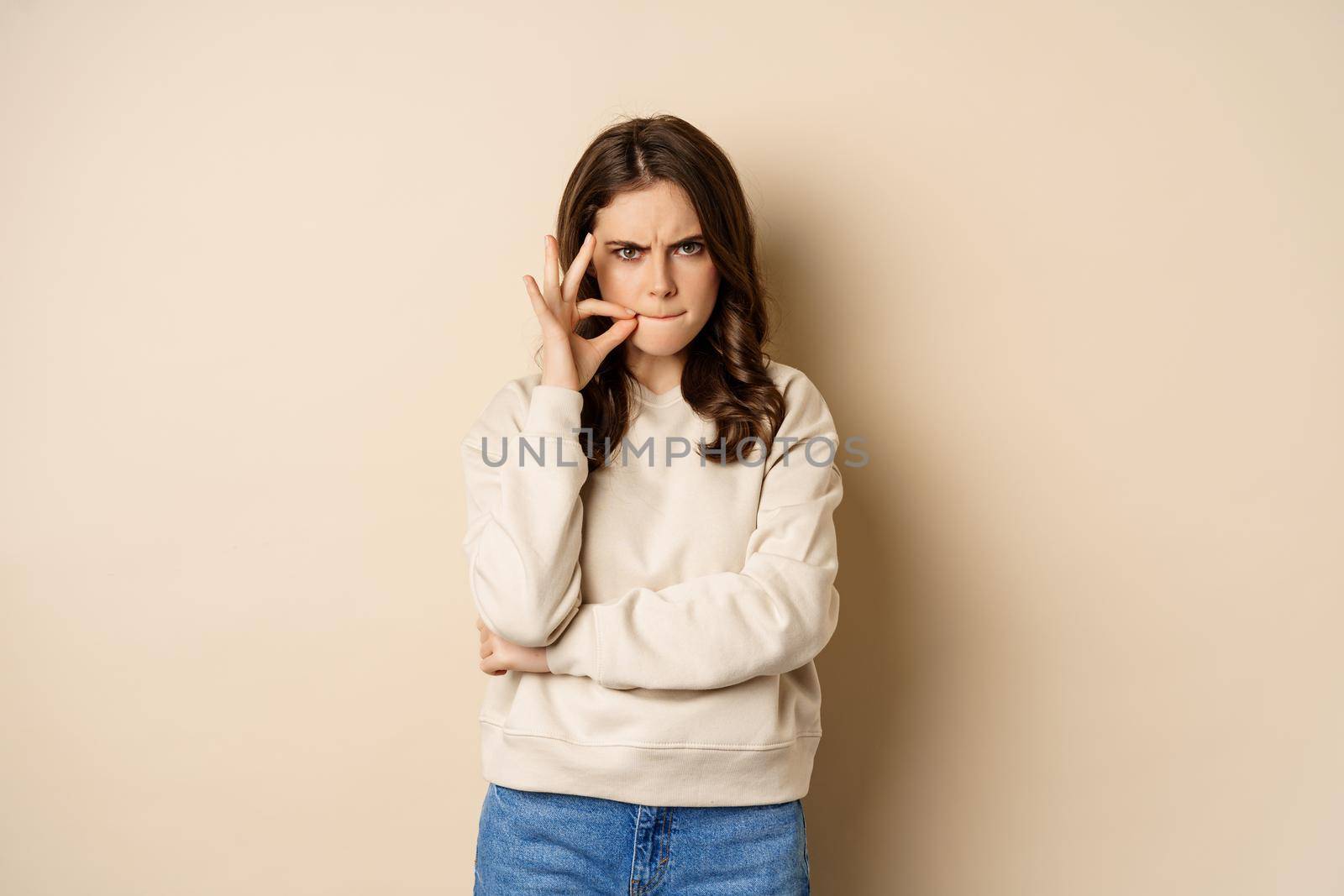 Angry woman shows mouth zipper, seal lips gesture, standing over beige background, taboo sign.