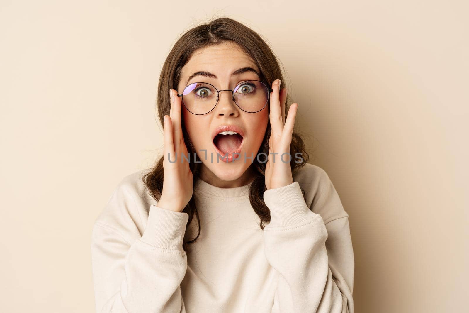Close up portrait of cute woman in glasses looking impressed, reacting amazed at smth she sees, standing over beige background.