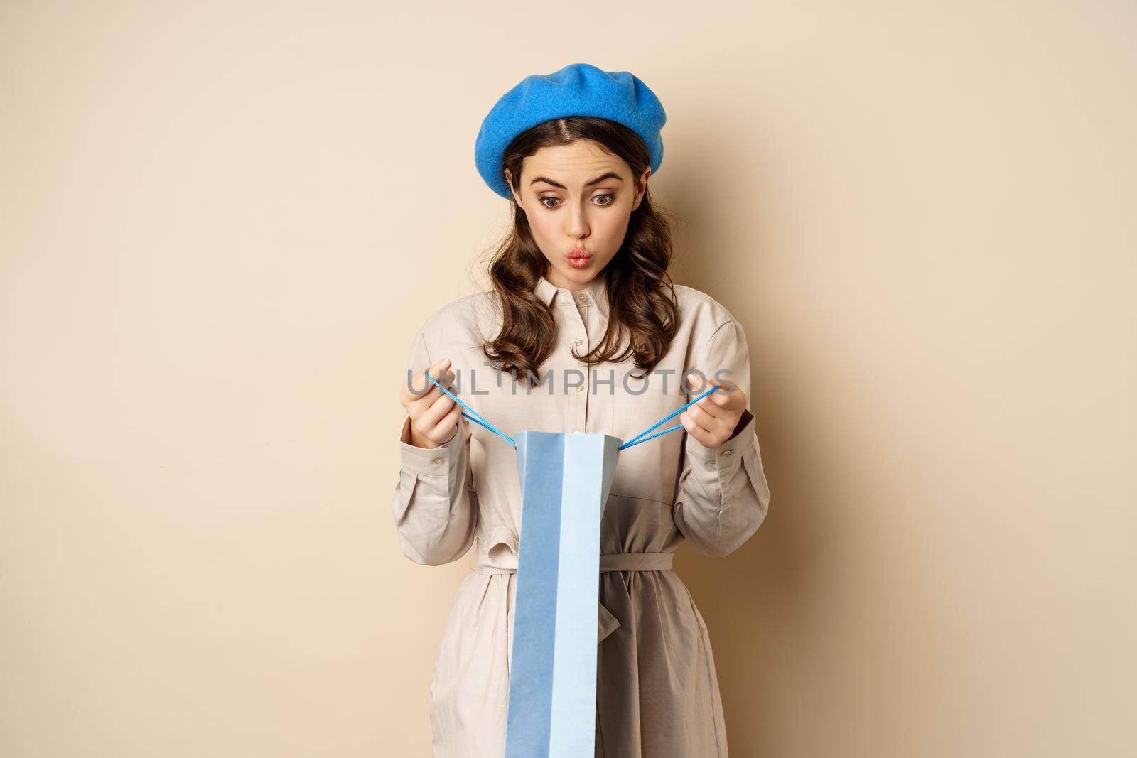 Image of girl looking surprised after opening gift bag, looking at present amazed, standing in stylish outfit over beige background by Benzoix