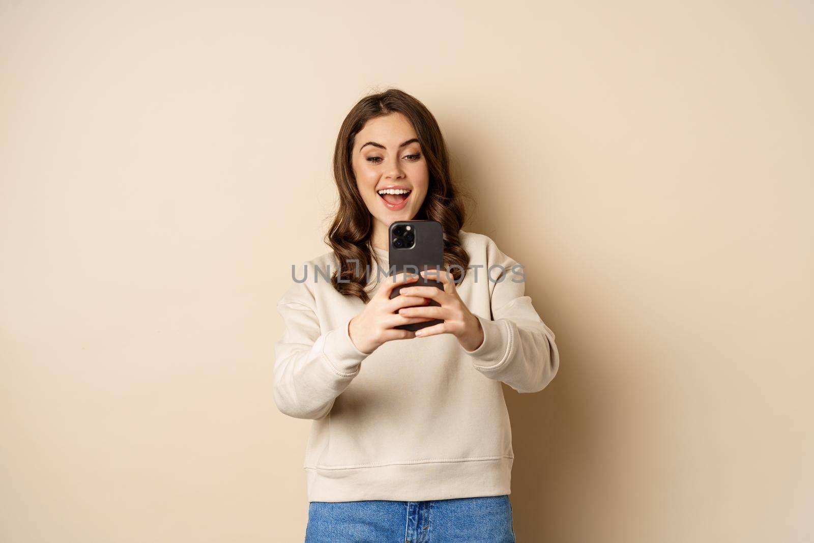 Happy woman recording video, shooting photo on smartphone camera and smiling, standing over beige background.