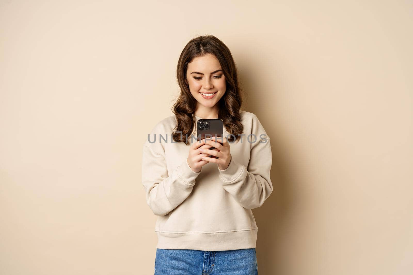 People and cellular technology. Beautiful stylish woman using mobile phone, smartphone app, smiling and looking at screen, standing over beige background by Benzoix