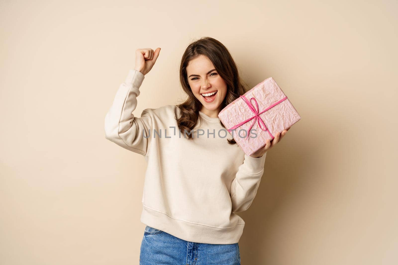 Happy smiling woman holding gift and triumphing, raising hand up joyfully, receive present on holiday, standing over beige background.
