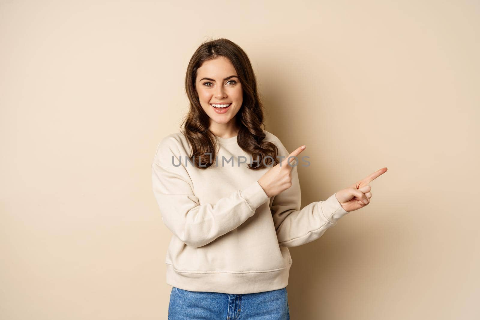 Smiling gorgeous woman pointing fingers right, inviting people, showing logo or banner, standing over beige background.
