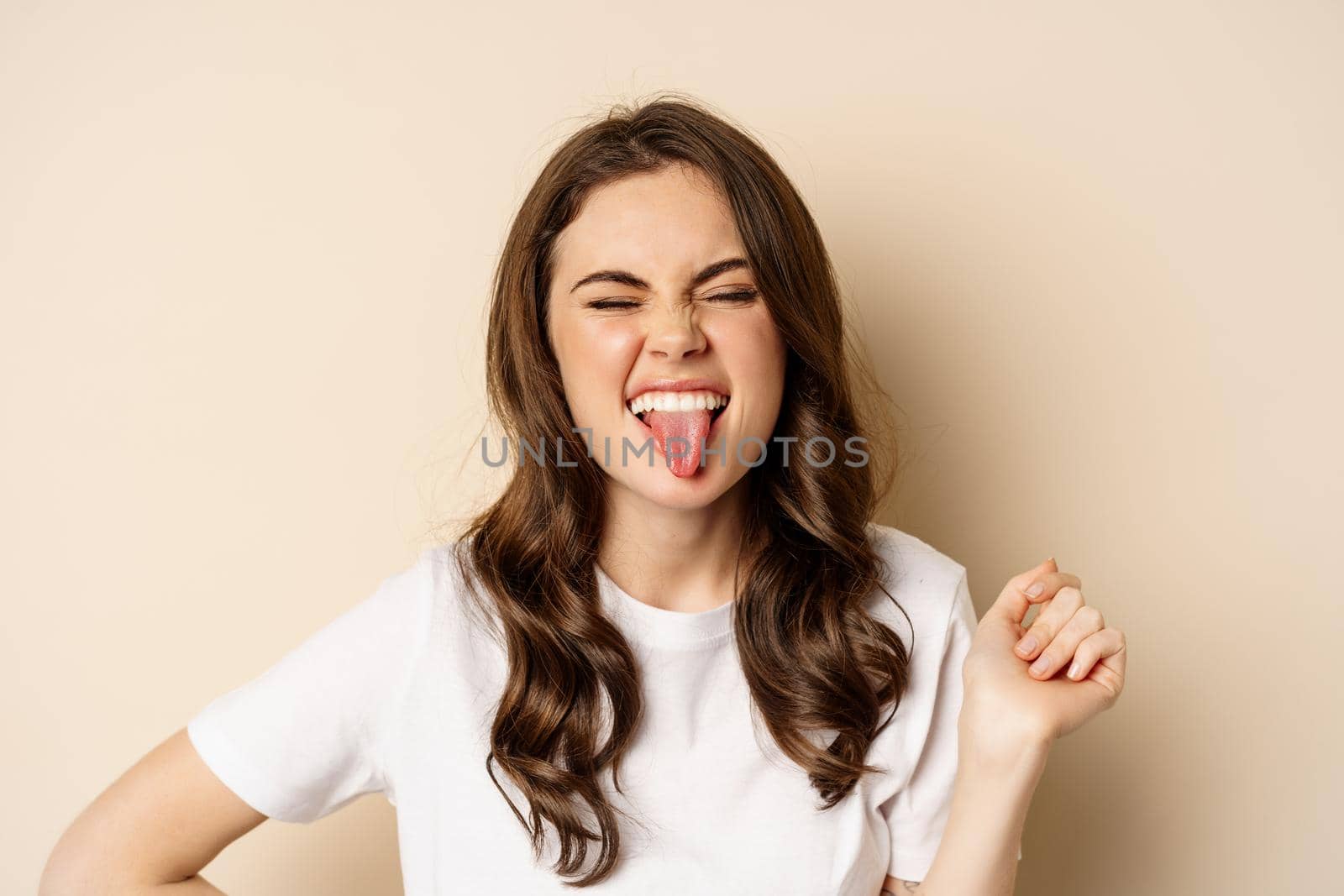 Close up of happy, carefree beautiful woman posing silly, showing tongue, having fun, standing in casual white t-shirt against beige background by Benzoix