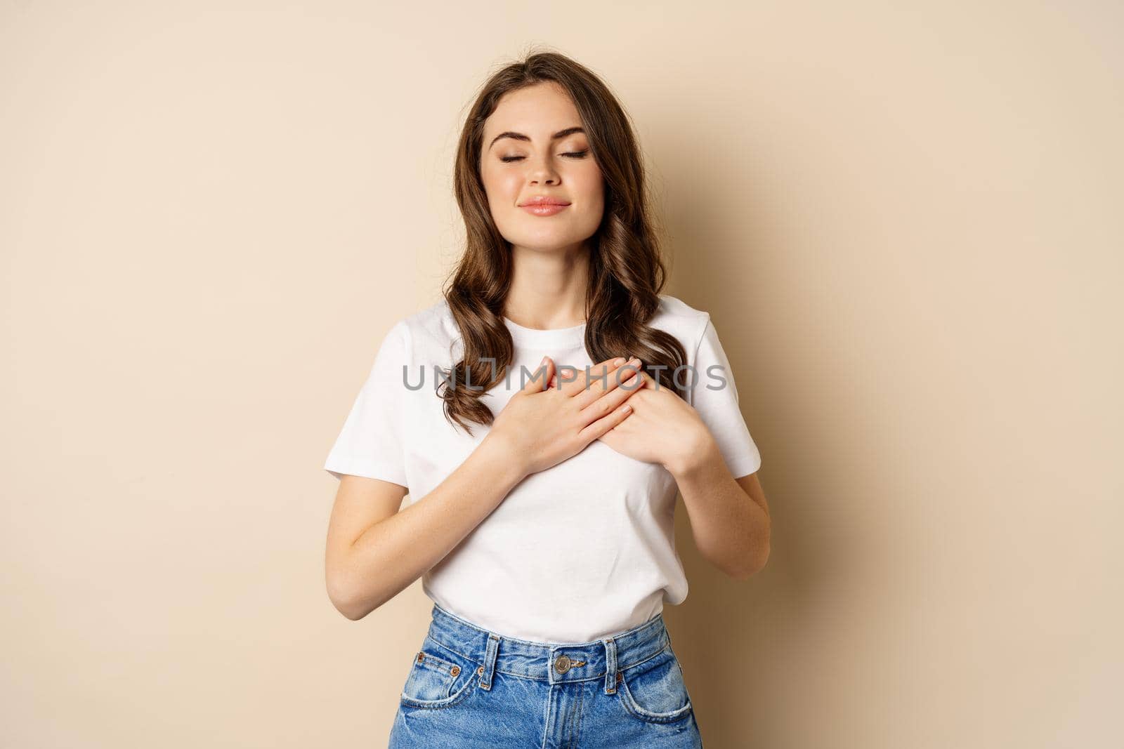 Portrait of young caucasian woman hold hands on heart and smiling, daydreaming, thinking of smth heartwarming, posing against beige background.