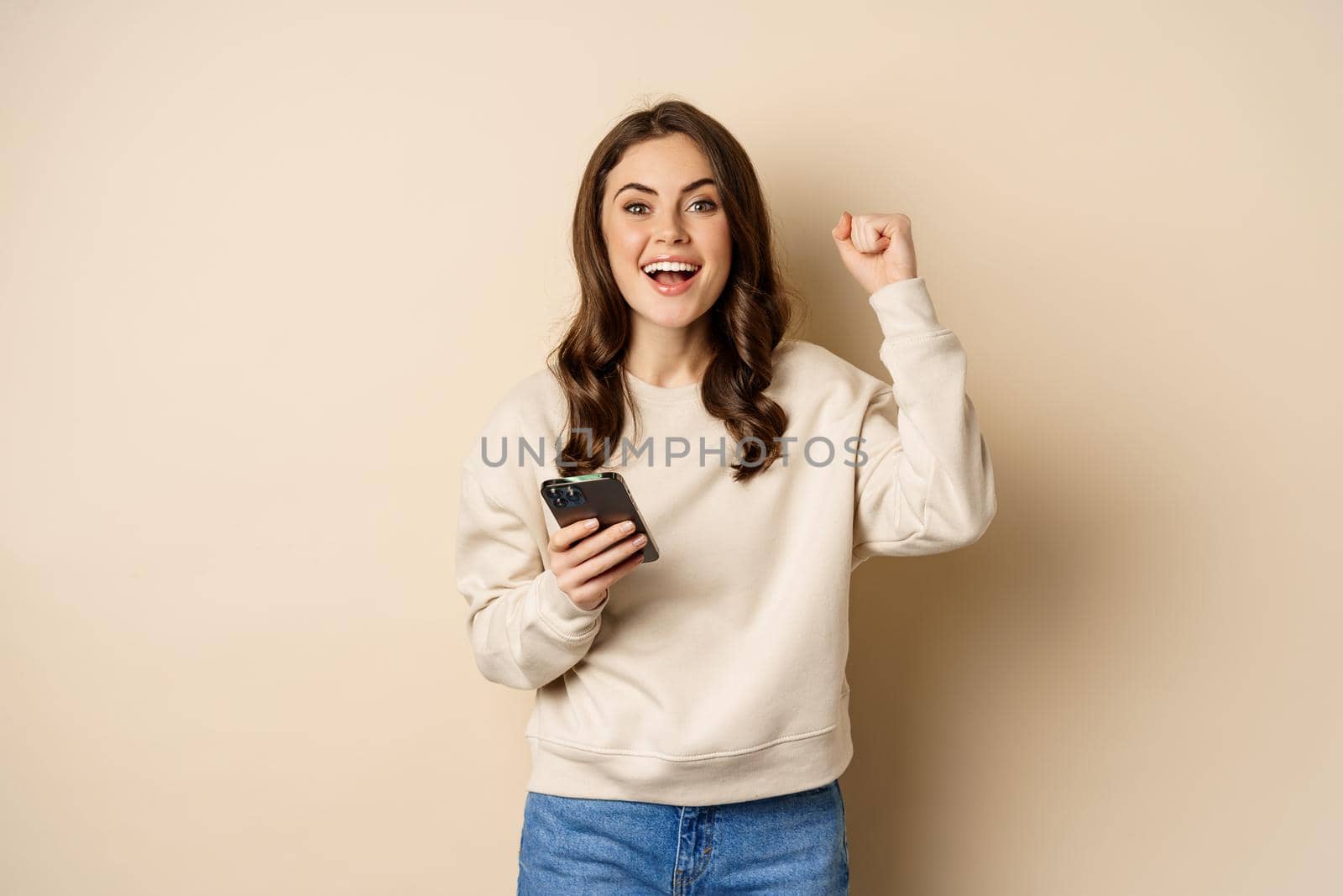 Happy brunette girl holding smartphone and cheering, winning, celebrating win on mobile phone app, standing over beige background by Benzoix