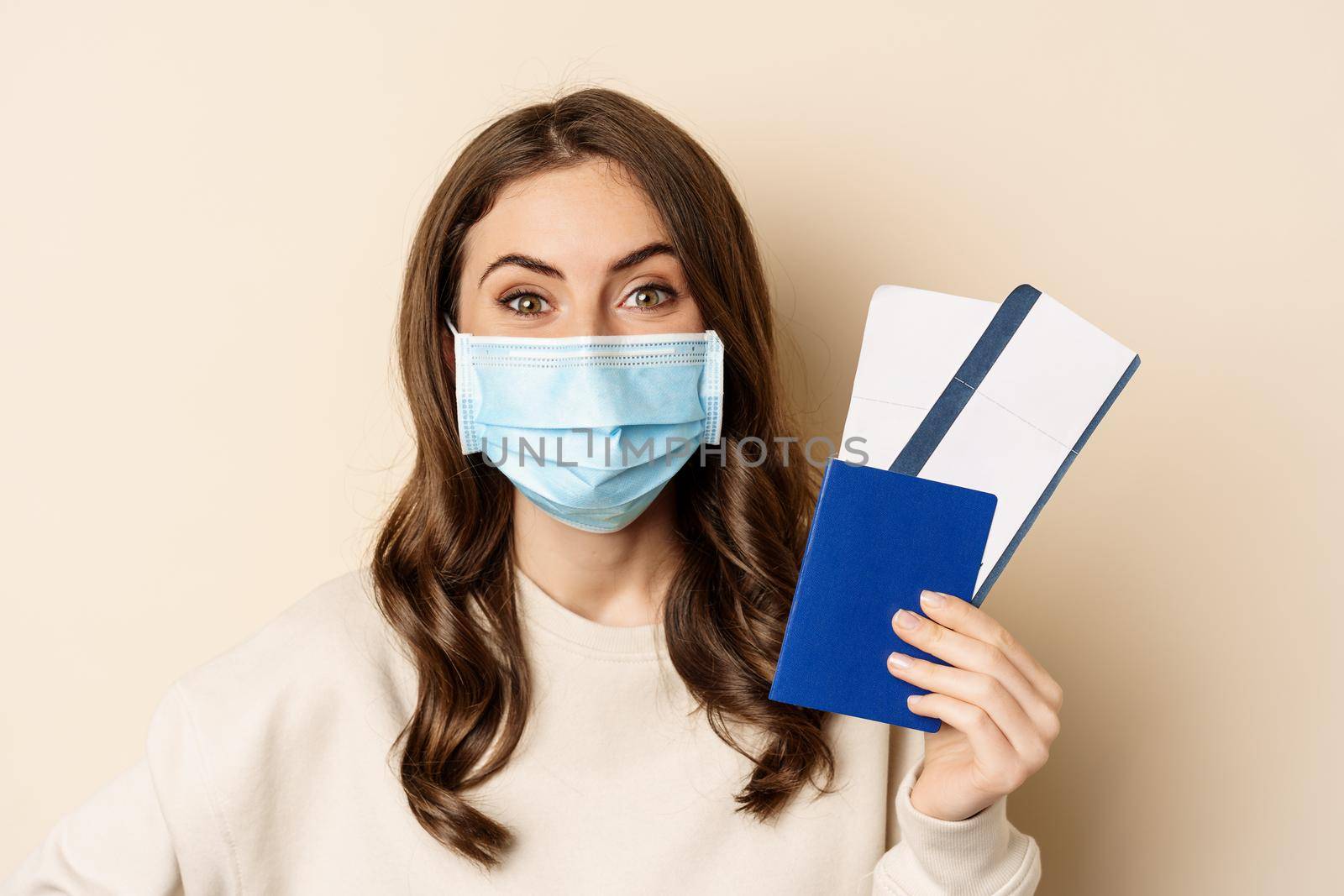 Travel and covid-19 pandemic. Close up portrait of smiling woman in medical mask, showing passport with two tickets, concept of tourism, beige background by Benzoix