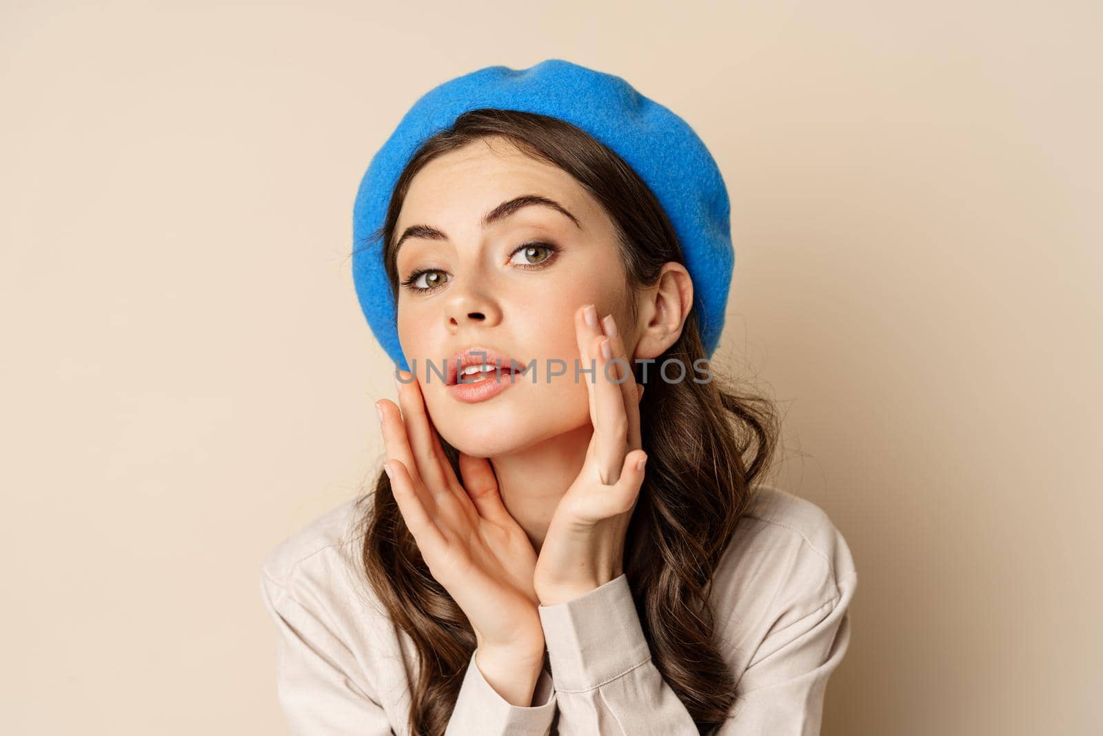 Close up portrait of young beatufiul woman looking in mirror and fixing her makeup, touching soft clear skin, standing over beige background by Benzoix