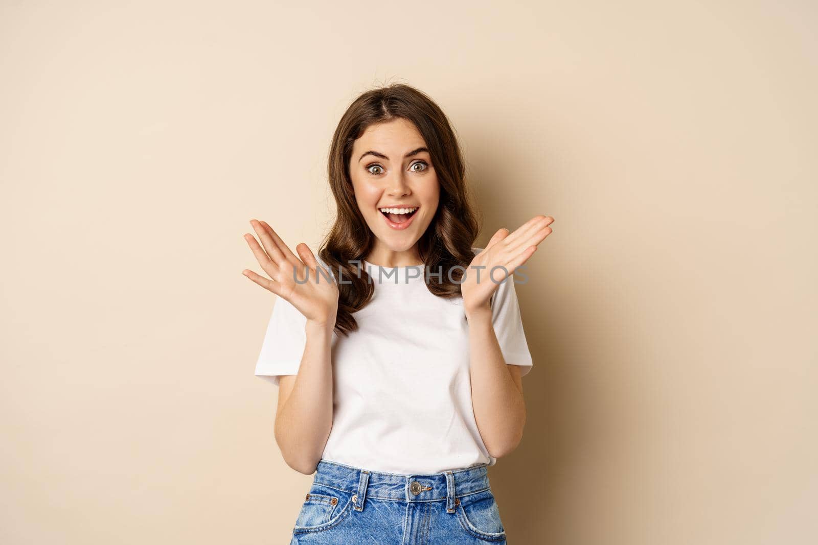 Portrait of happy young woman rejoicing, clapping hands and smiling, applause, standing over beige background.