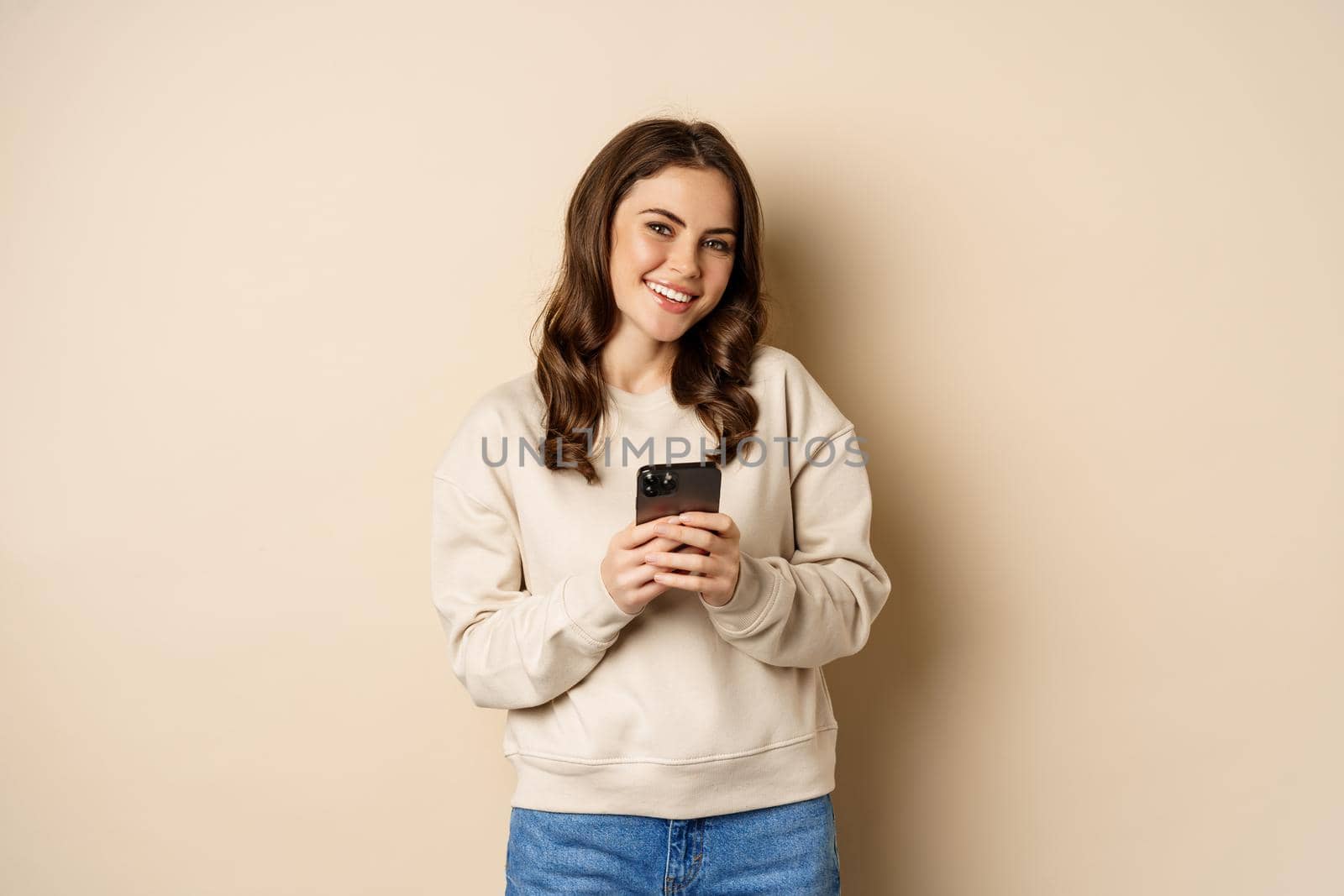 People and cellular technology. Beautiful stylish woman using mobile phone, smartphone app, smiling and looking at screen, standing over beige background.