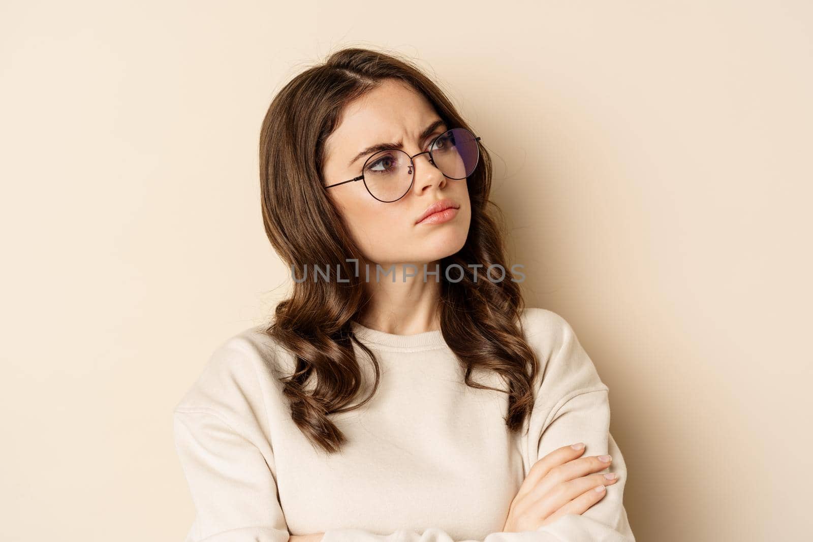 Close up portrait of complicated, troubled woman in glasses, frowning and looking displeased, standing over beige background by Benzoix