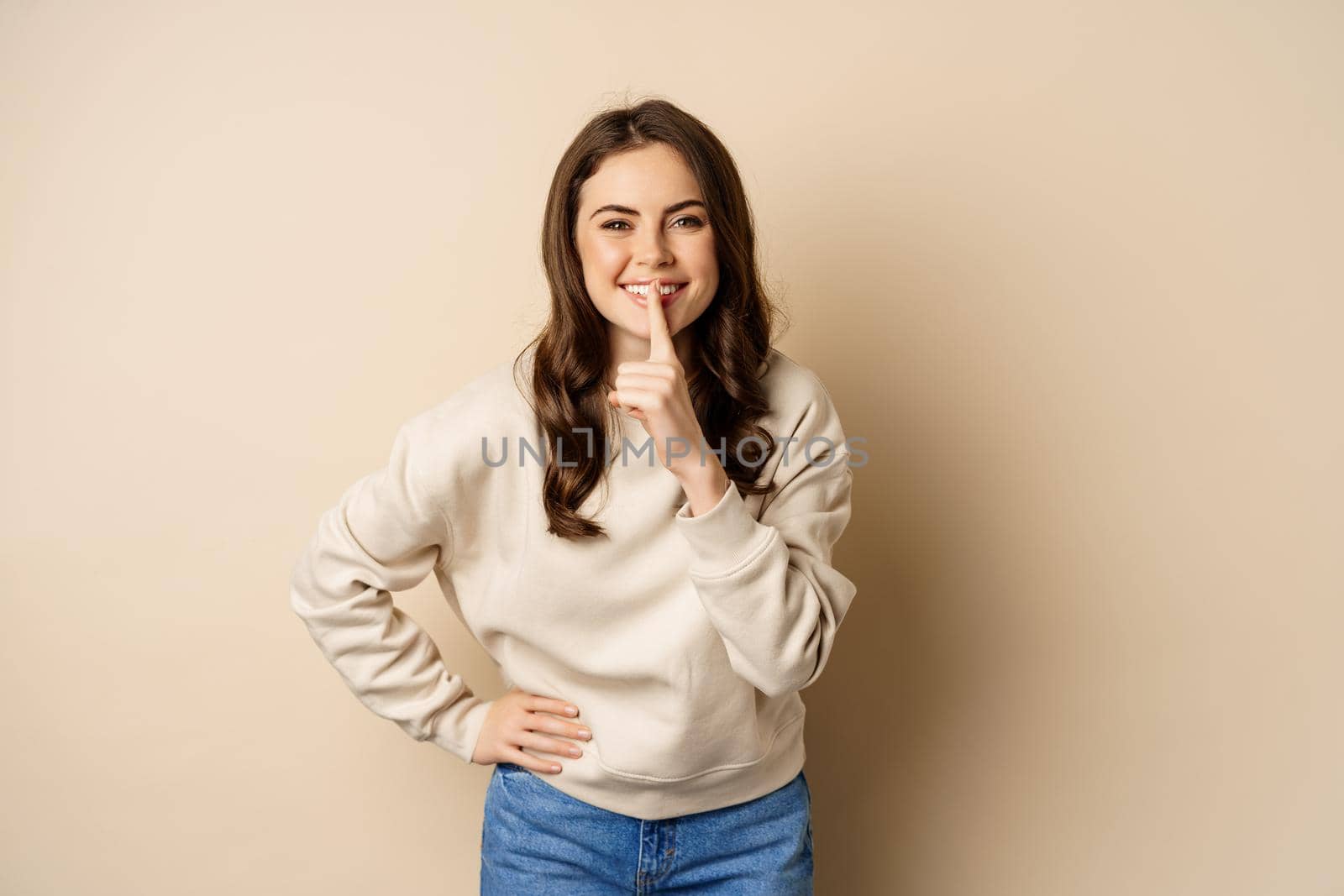 Happy coquettish girl say shh, shushing, keep secret, be quiet gesture, smiling at camera, standing over beige background by Benzoix