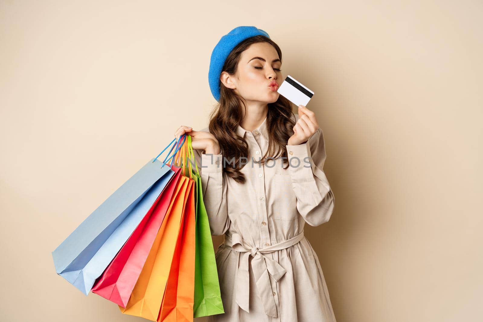 Portrait of cute happy girl kissing her credit card after buying gifts, holding shopping bags, standing over beige background by Benzoix