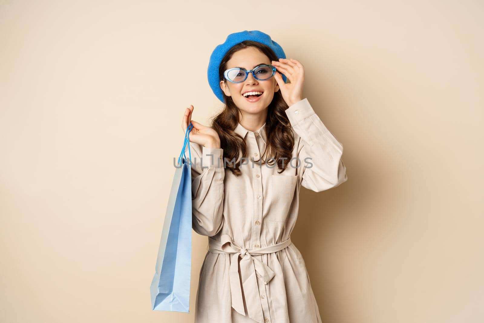 Stylish happy girl on shopping. Portrait of modern woman with shop bag, laughing and smiling satisfied, buying herself gift, standing over beige background.