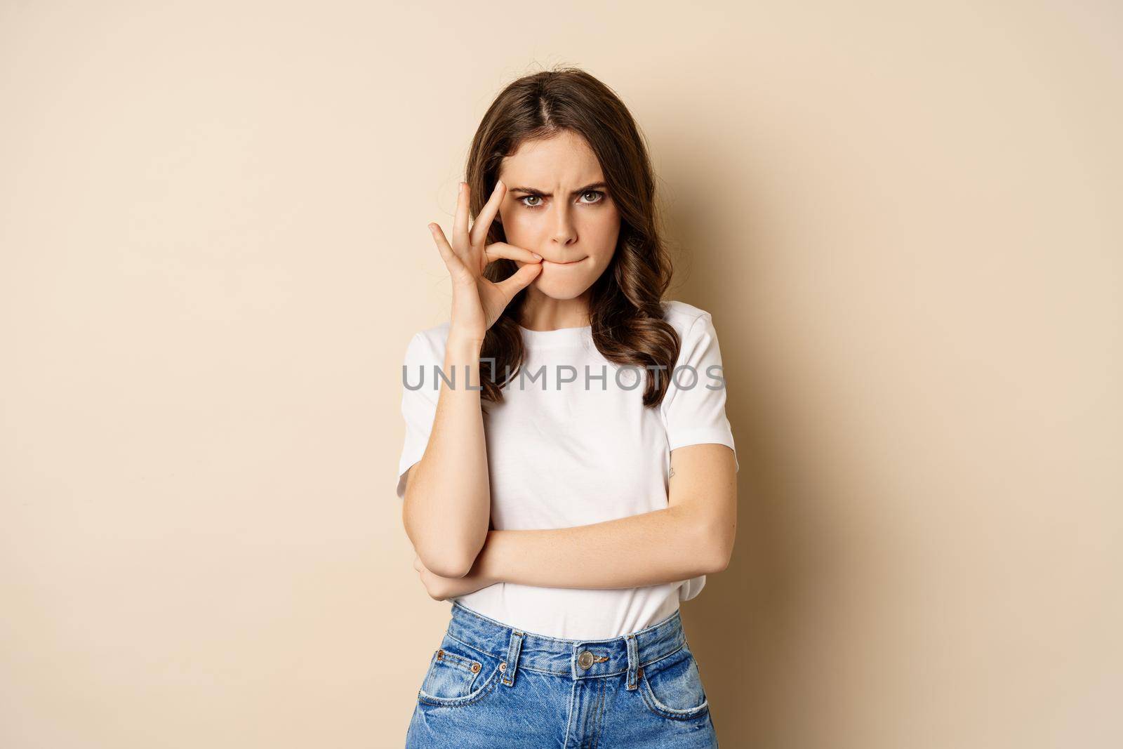 Young serious woman seal lips, showing zip mouth gesture, standing over beige background.