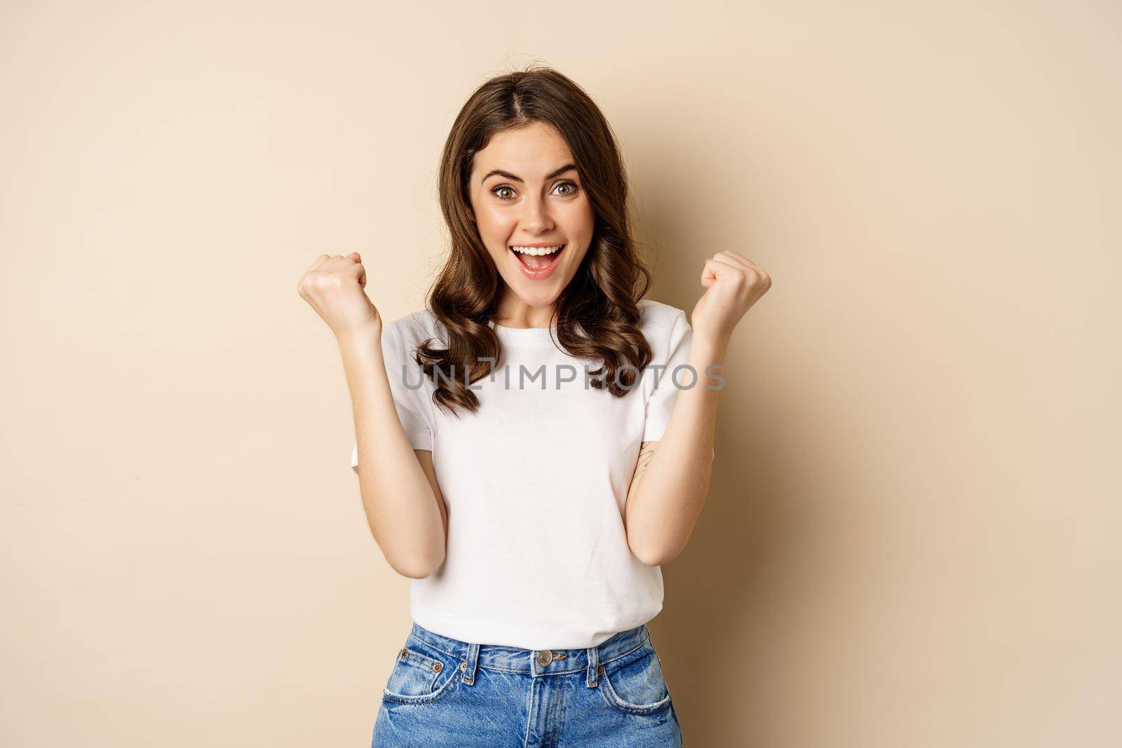 Enthusiastic woman winning and fist pump, celebrating victory, smiling excited, standing over beige background.