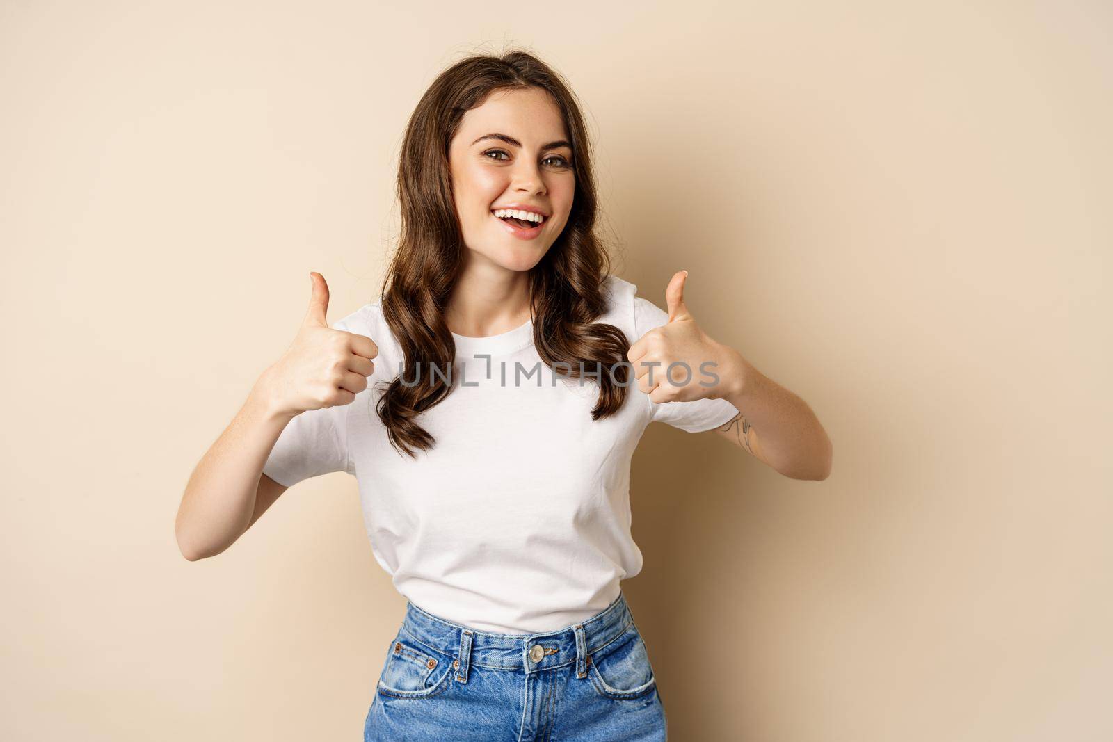 Excellent work. Happy young woman showing thumbs up in approval and smiling, standing in t-shrit over beige background by Benzoix