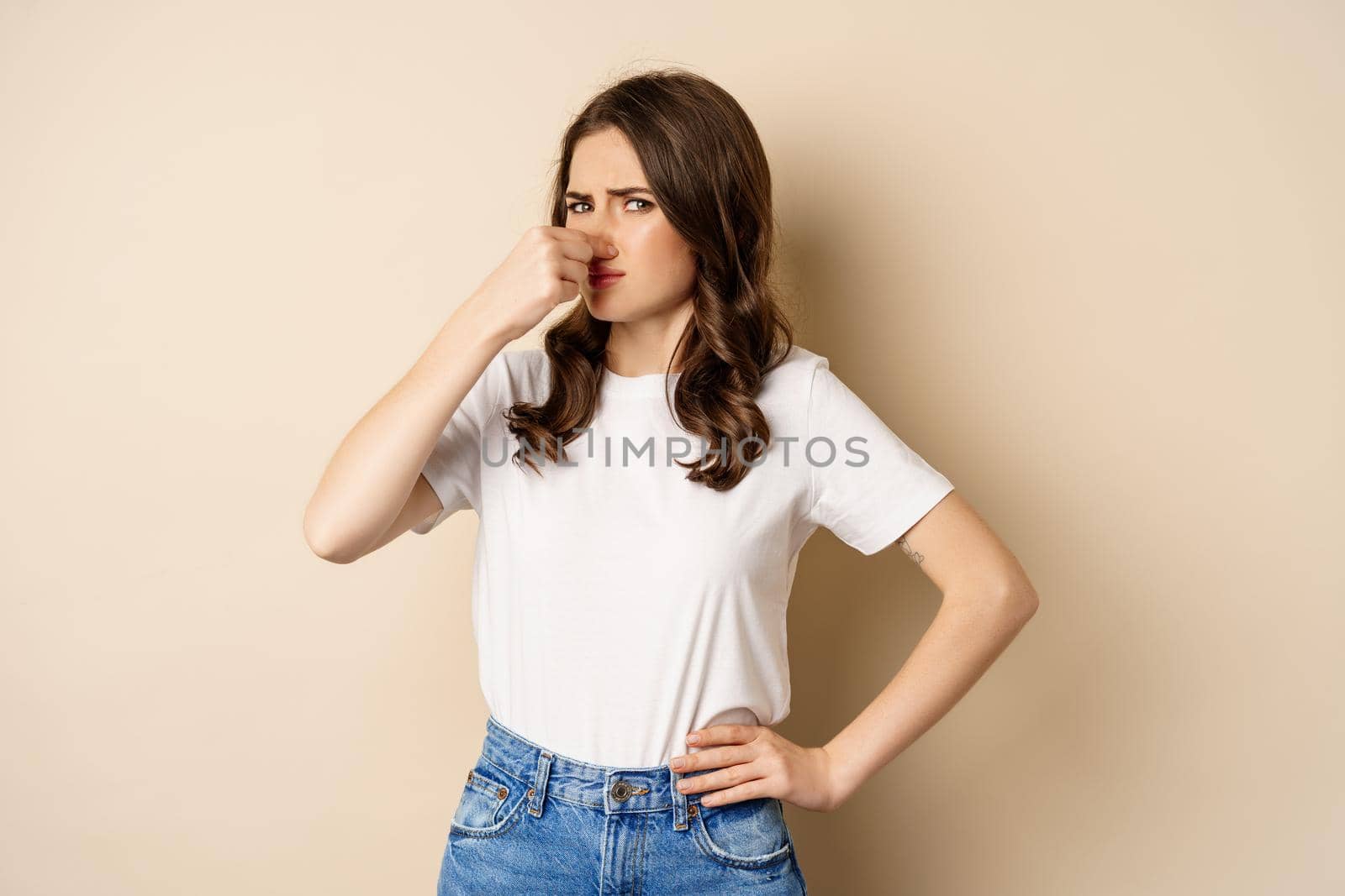 Woman shut nose from disgusting smell, standing over beige background.