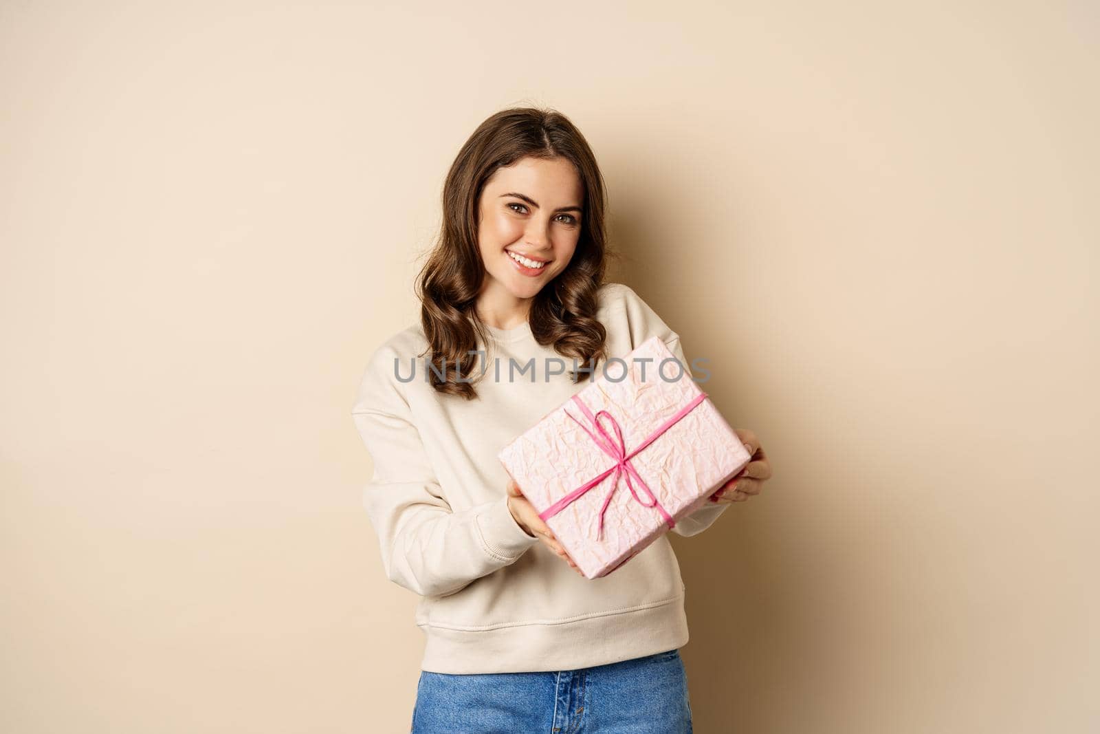 Beautiful girl posing with gift and smiling, showing wrapped present box, standing in sweater over beige background. concept of holidays.