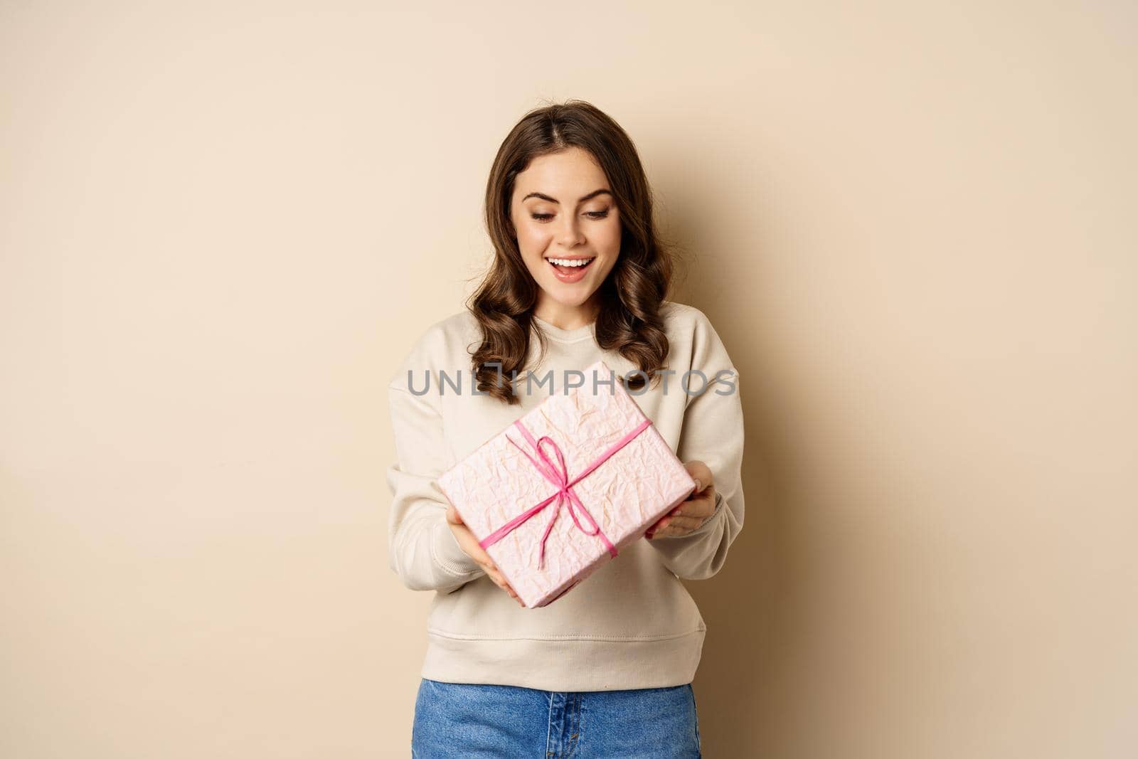 Excited beautiful girl with pink wrapped gift box, receive presents, standing over beige background.