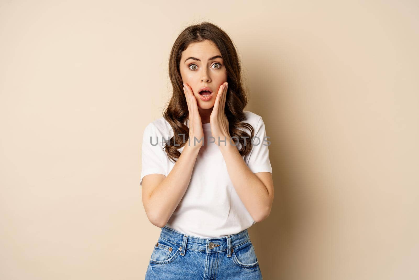 Portrait of stylish young woman gasping, looking surprised and amazed, impressed by smth, standing in t-shirt and jeans over beige background.