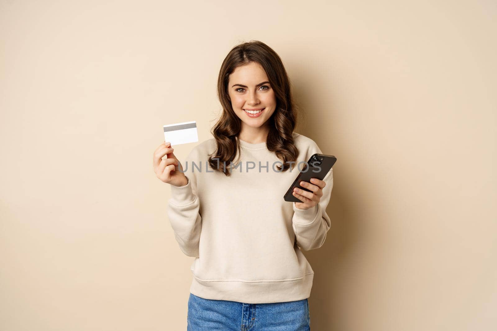 Online shopping. Smiling brunette woman showng credit card, using smartphone app, standing over beige background.