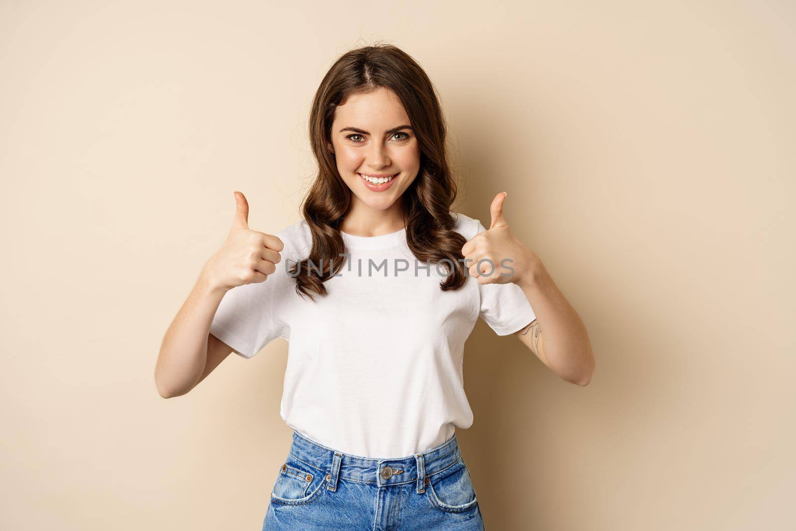 Excellent work. Happy young woman showing thumbs up in approval and smiling, standing in t-shrit over beige background by Benzoix