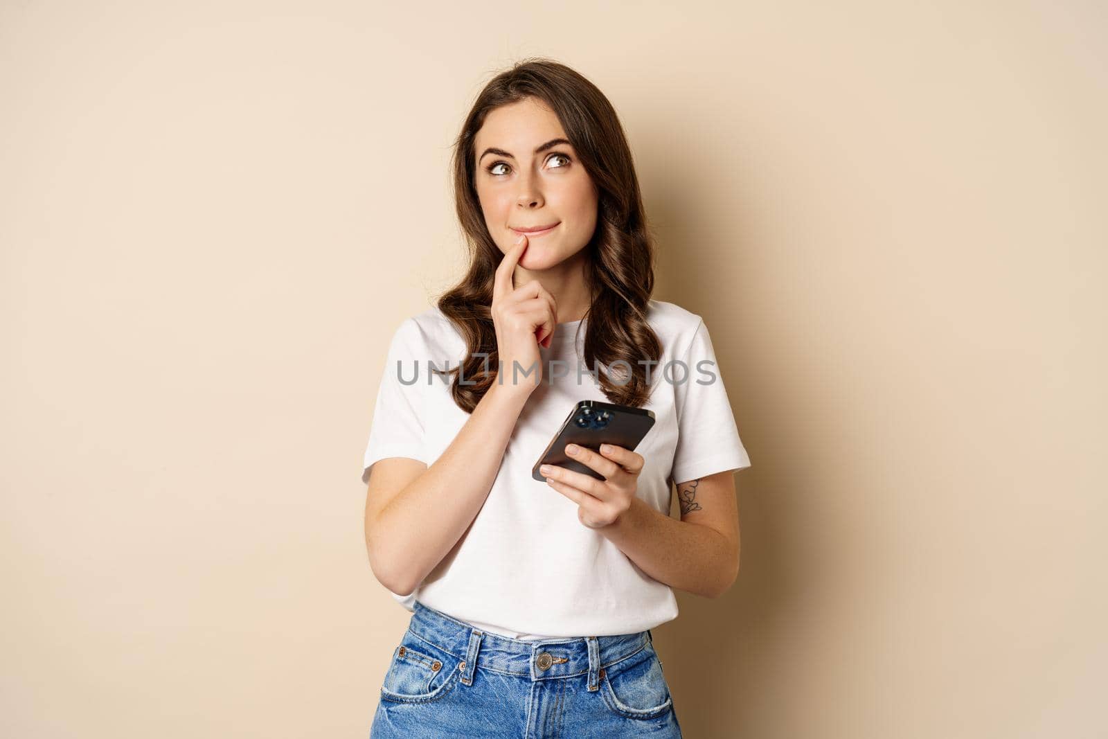 Girl thinks, holds mobile phone, looks up thoughtful, stands over beige background.