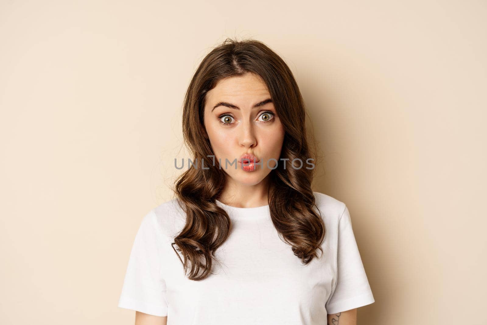 Close up portrait of surprised and excited brunette girl looking amazed, reacting impressed and excited, standing against beige background by Benzoix