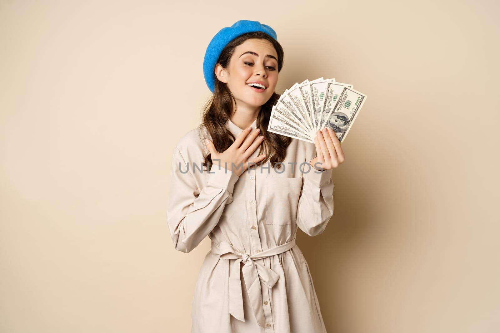 Stylish young woman holding money, cash dollars, smiling and posing satisfied, going on shopping, standing over beige background.