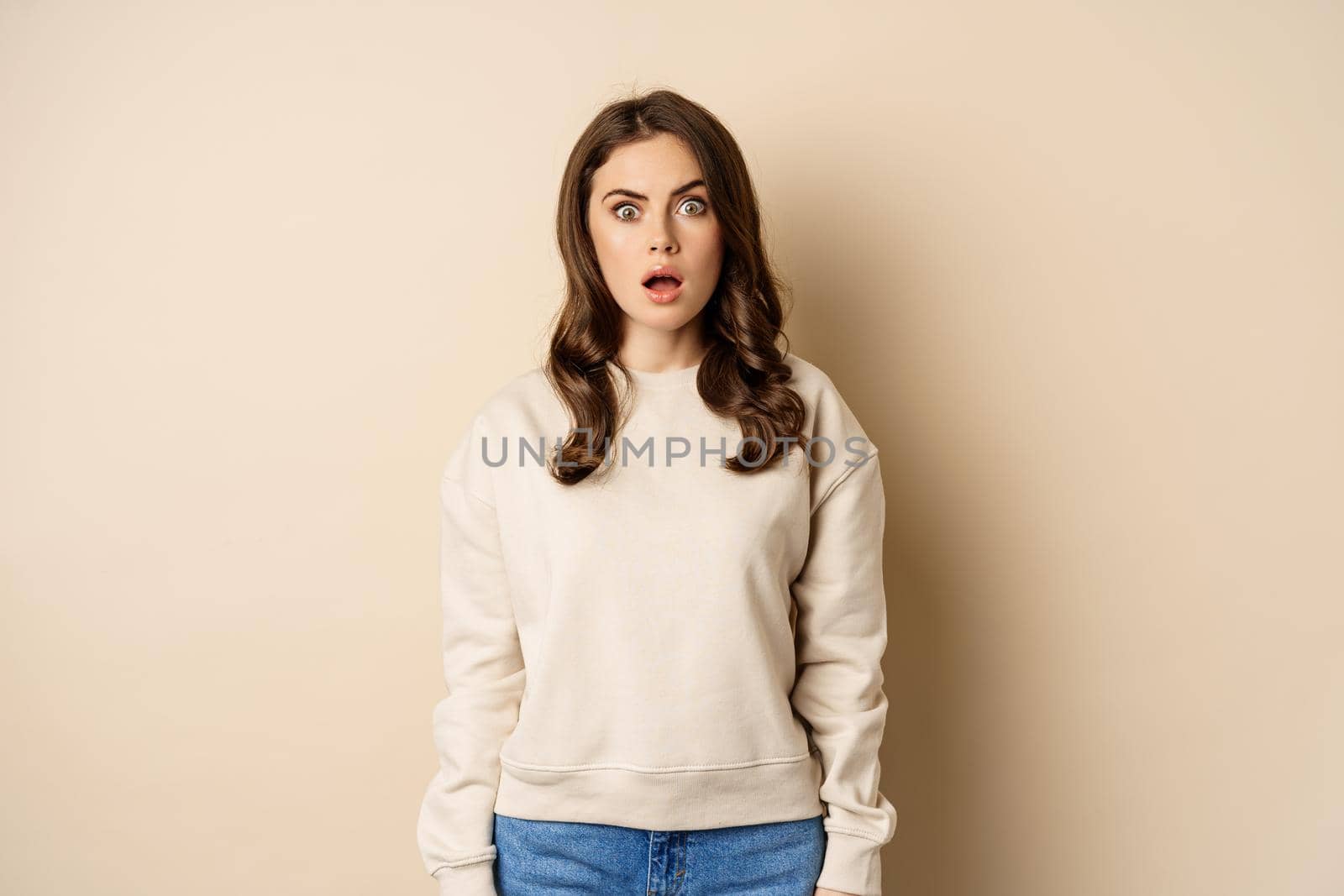 Portrait of shocked brunette woman drop jaw, gasping and staring speechless at camera, beige background by Benzoix