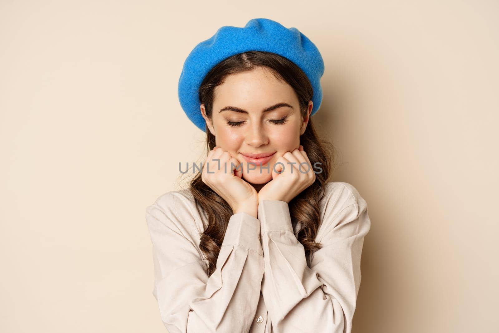 Close up portrait of feminine young woman in trendy french hat, posing cute and romantic, making silly coquettish face and tender smile, beige background.