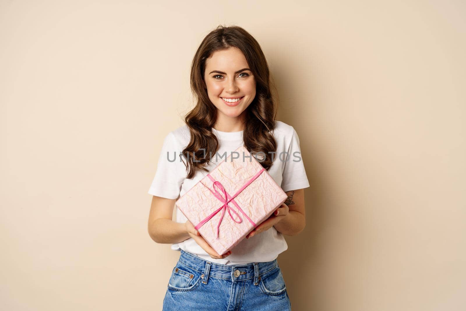 Celebration and holidays concept. Happy young woman holding gift wrapped in pink box, receive present, looking amazed and surprised, standing over beige background.