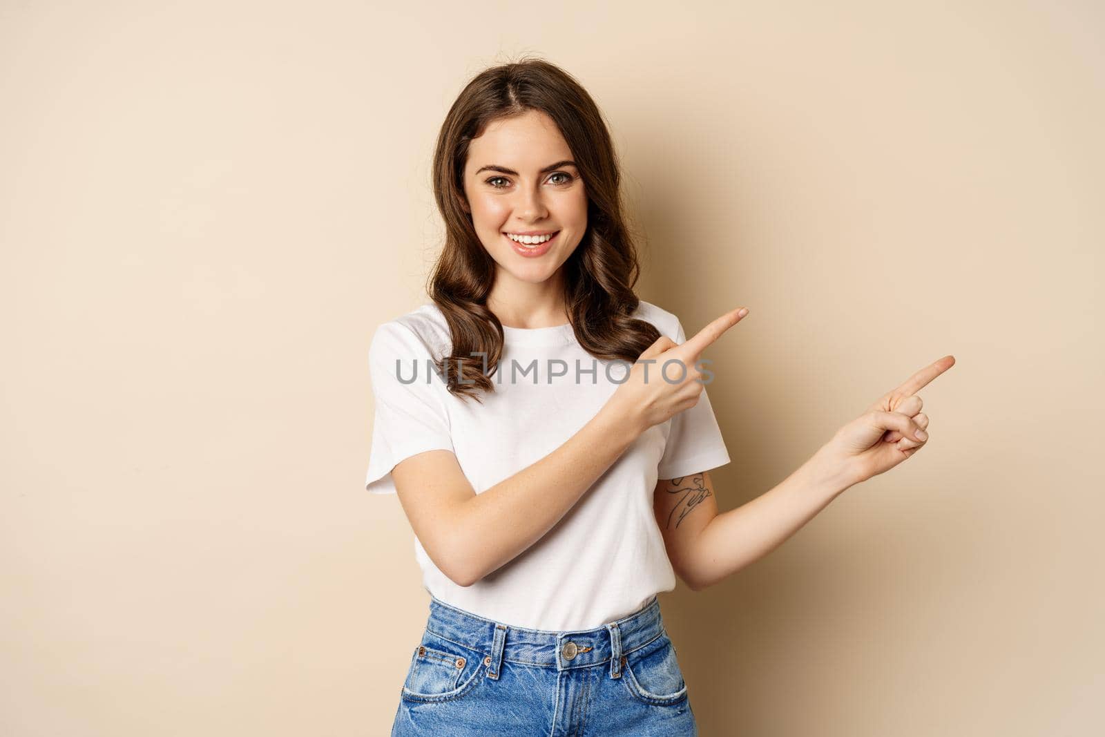 Smiling brunette woman in t-shirt, pointing fingers right, showing promo offer or advertisement, demonstrating banner, standing against beige background by Benzoix