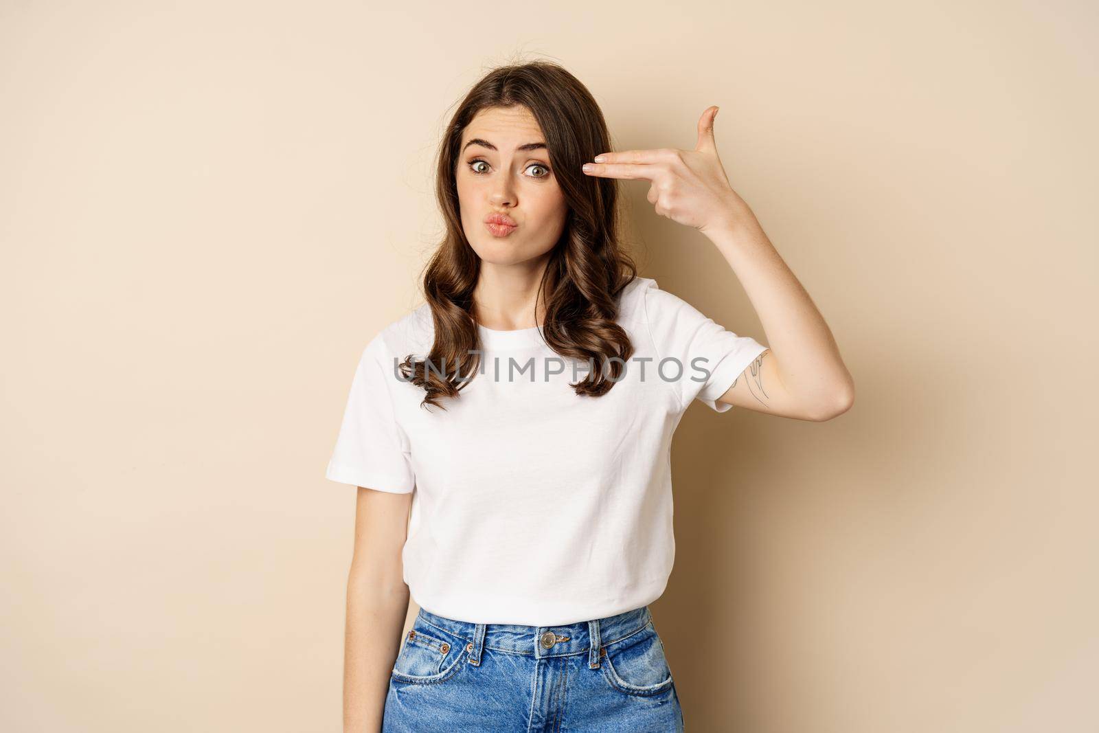 Young woman showing finger pistol sign near head and looking annoyed, sick and tired, standing over beige background.