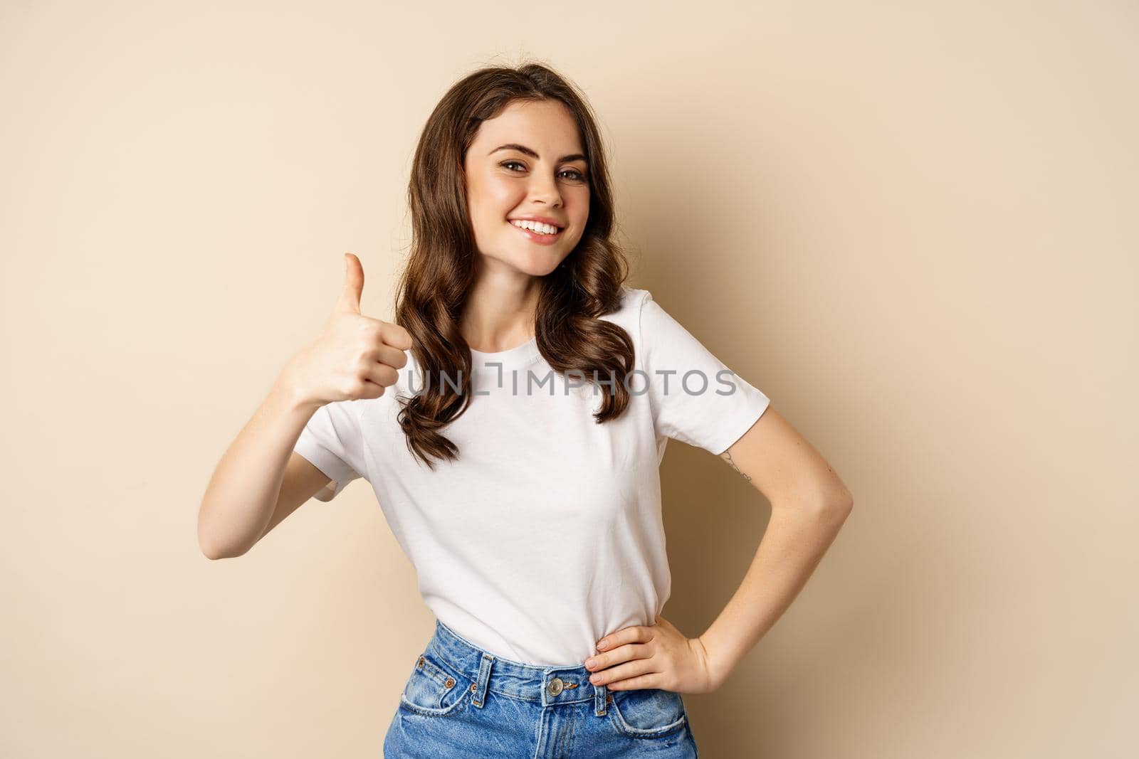 Portrait of modern young woman showing thumbs up, like and approve, smiling pleased, recommending company or website, standing over beige background.