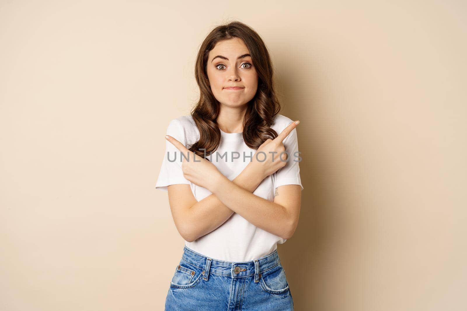 Beautiful girl choosing between two options, pointing sideways and looking clueless, standing over beige background.