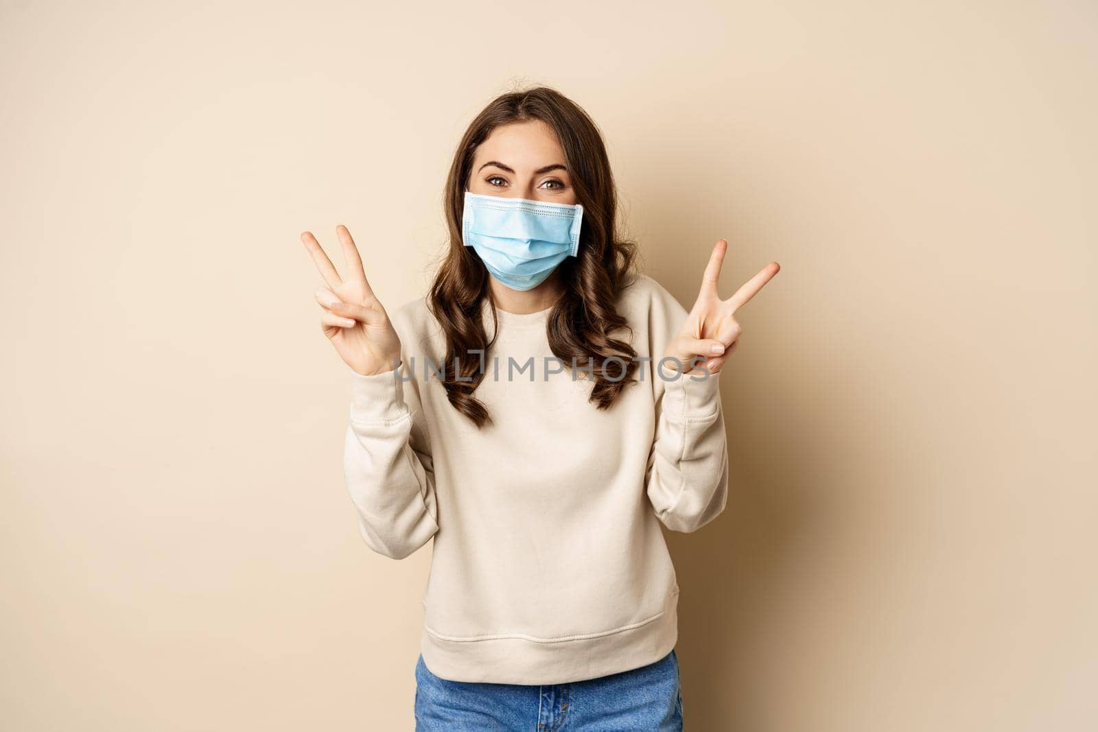 Covid-19, pandemic and quarantine concept. Beautiful adult woman in face medical mask, showing peace, v-sign gesture, standing over beige background.