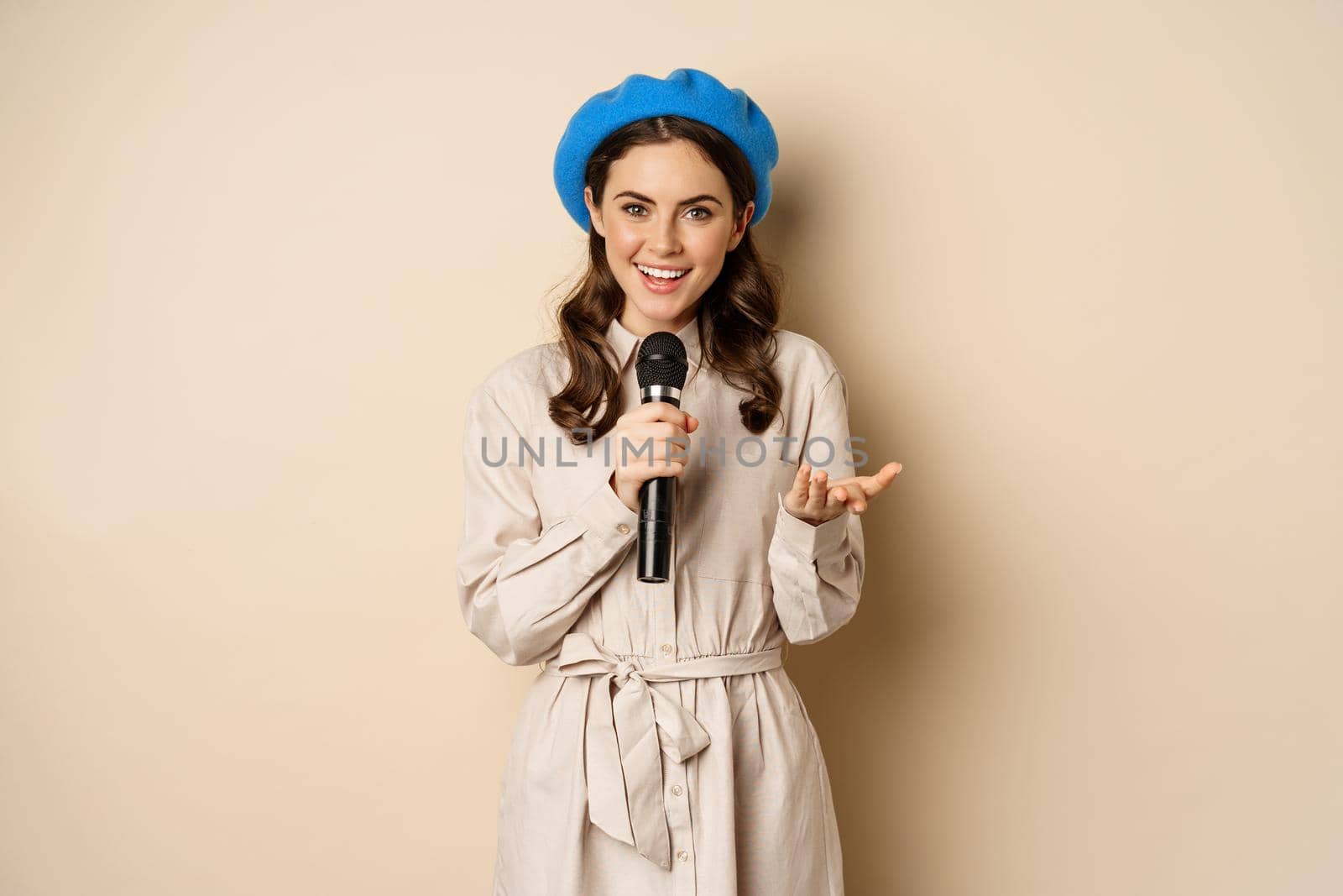 Portrait of happy stylish woman performing, singing with microphone, posing against beige background by Benzoix