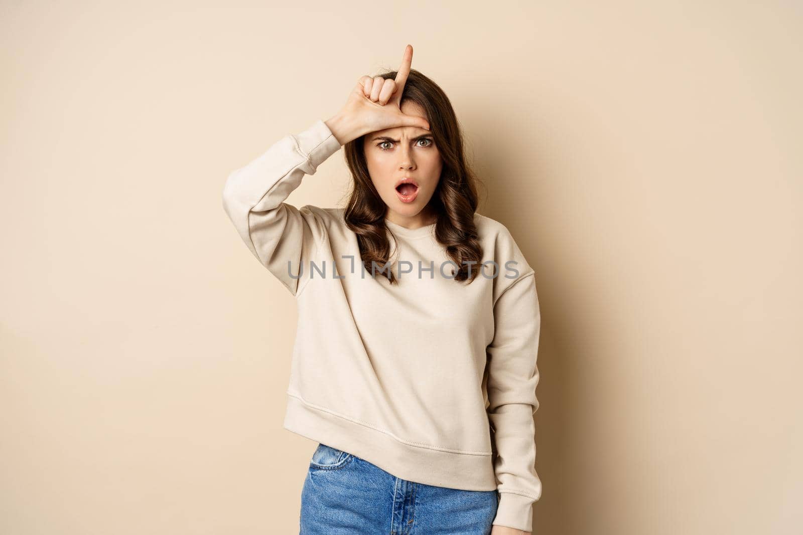 Woman showing loser sign on forehead, L word, standing over beige background by Benzoix