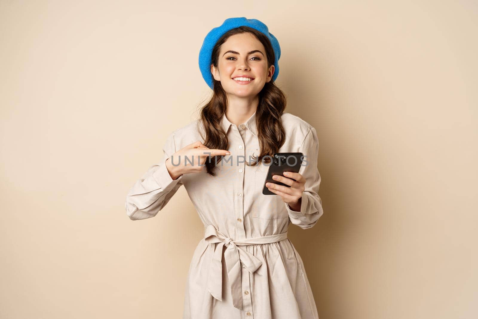 Portrait of stylish modern woman in outerwear, pointing at mobile phone screen and looking happy, smiling pleased, standing over beige background.