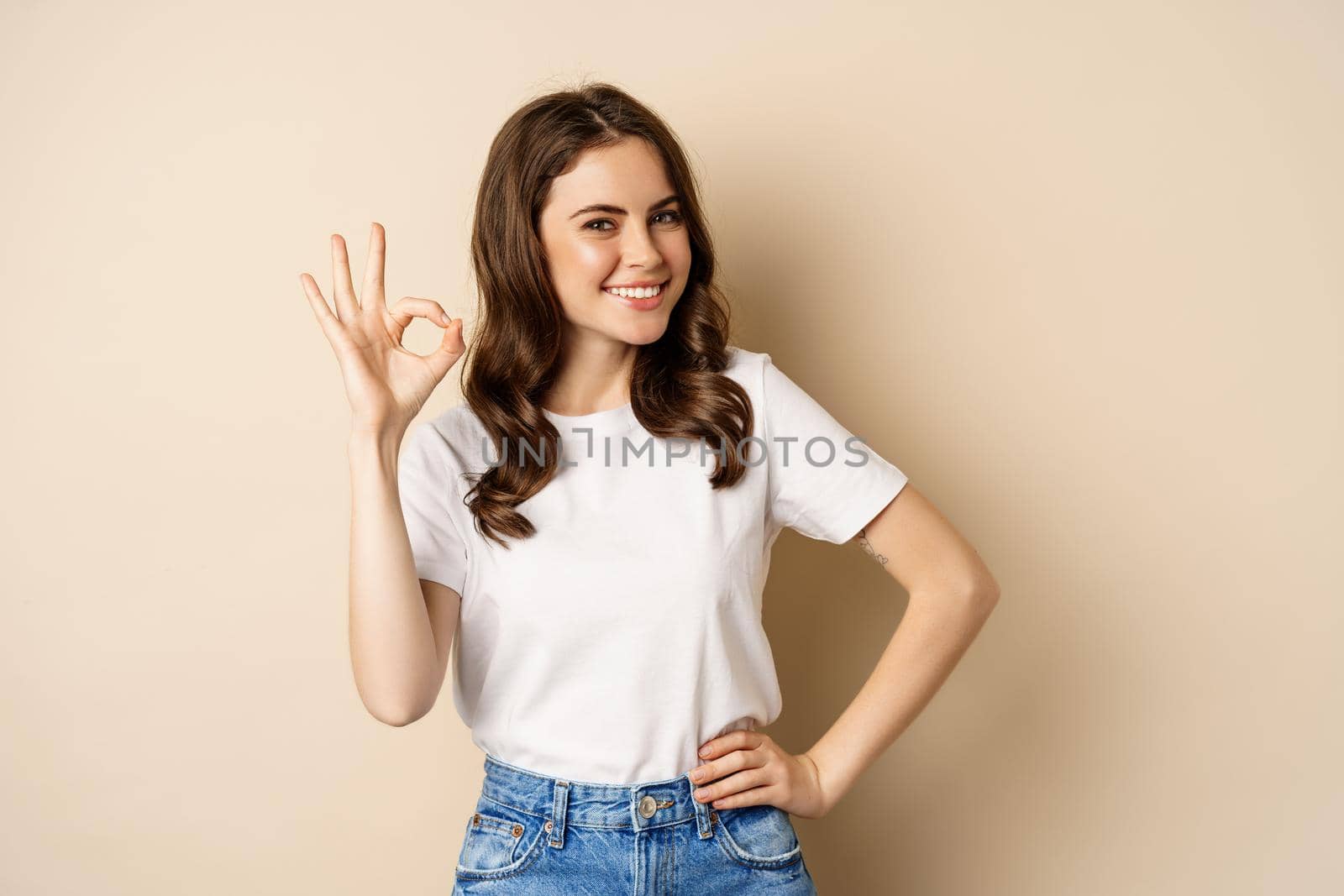 Stylish young woman smiling satisfied, showing okay, ok sign in approval, recommending, standing over beige background by Benzoix