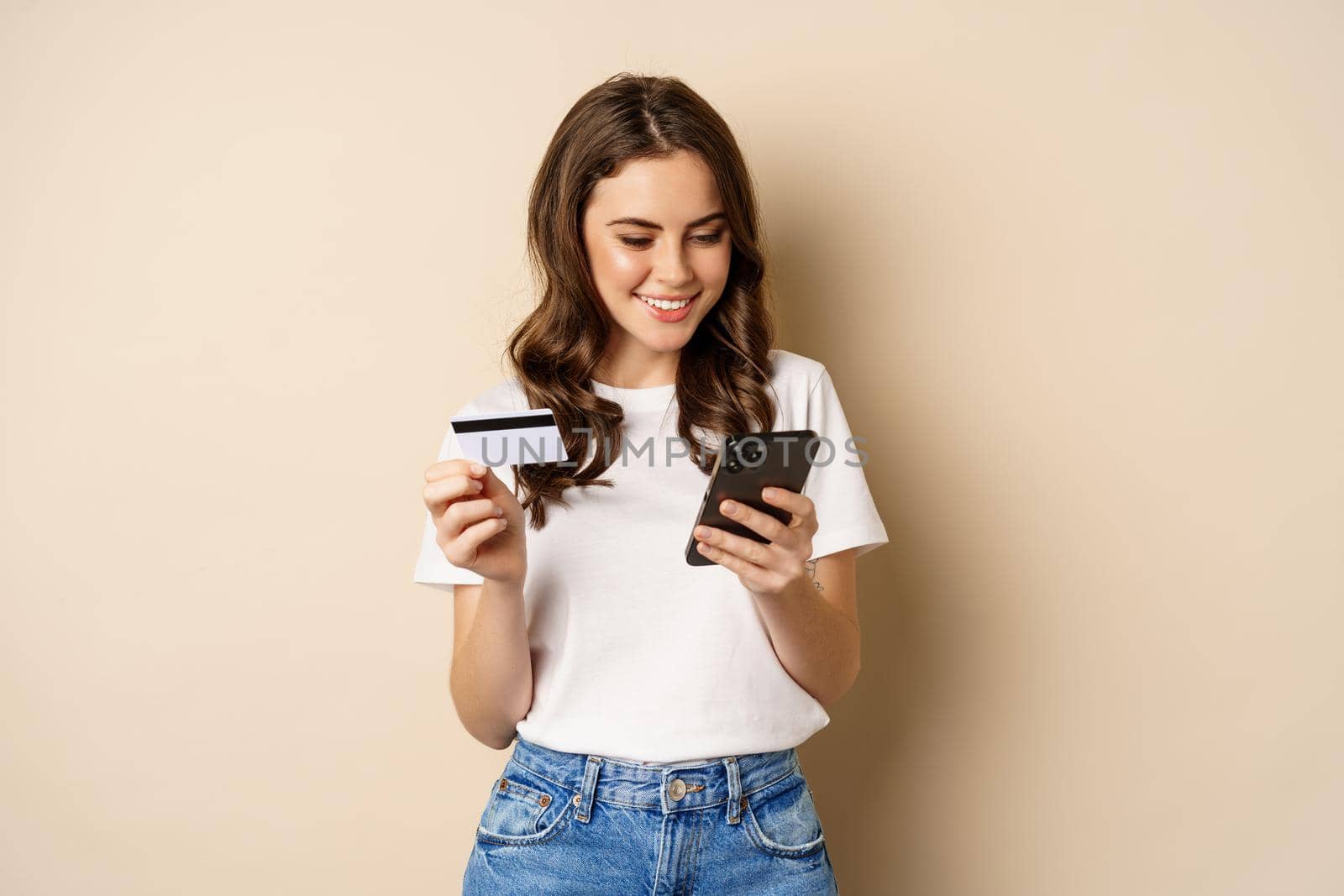 Image of young happy woman paying online, holding smartphone and credit card, enter info in application on mobile phone, standing against beige background.