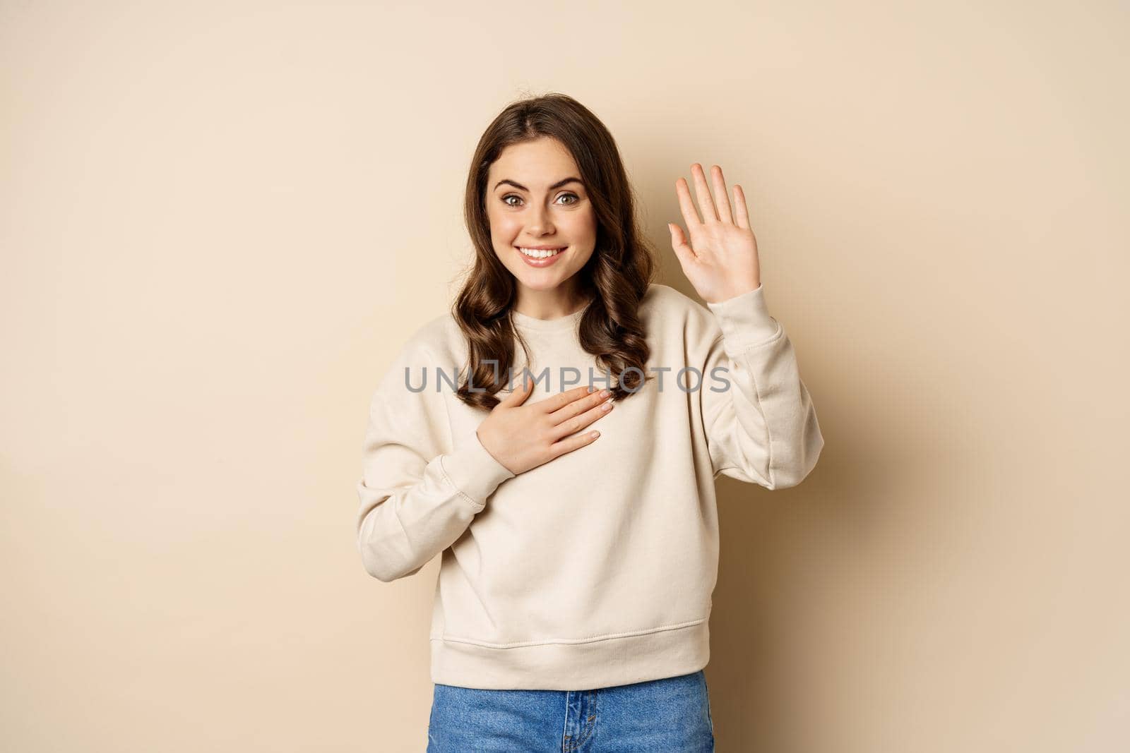 Cute modest girl smiling, raising finger, introduce herself, saying hello, standing over beige background.